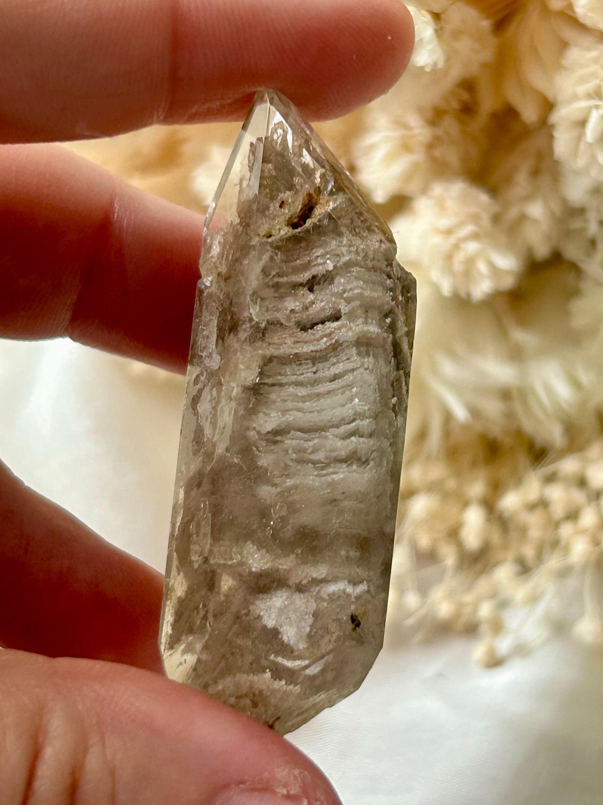 holding a Lodolite freeform crystal against an ivory satin backdrop with dried cream flowers; soft natural light highlights internal inclusions.