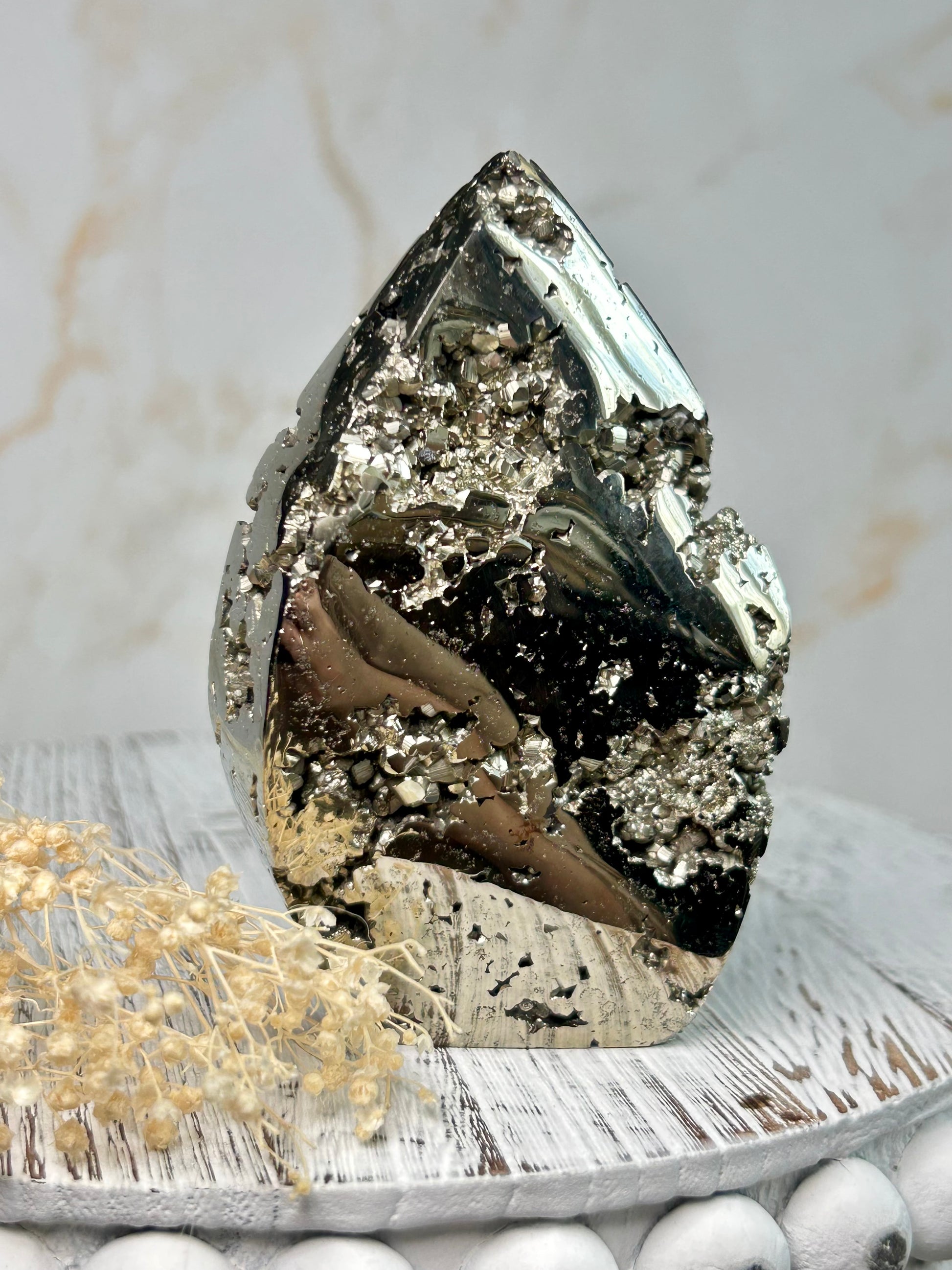 Golden pyrite flame crystal polished into flame shape, shimmering metallic surface displayed on a distressed wooden board with cream dried flowers.