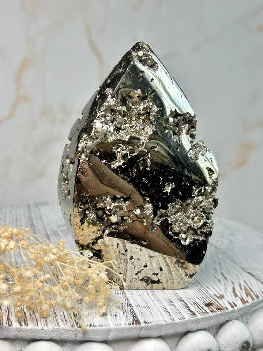 Golden pyrite flame crystal polished into flame shape, shimmering metallic surface displayed on a distressed wooden board with cream dried flowers.