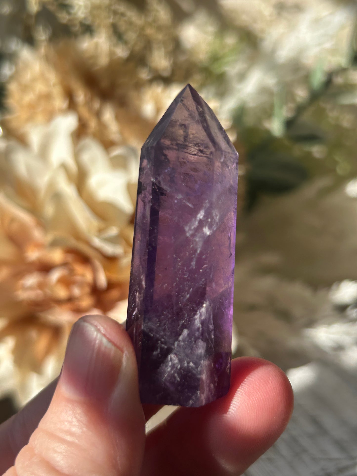 A hand holding a pointed purple amethyst crystal point against a blurred natural background of rocks and dried flowers.