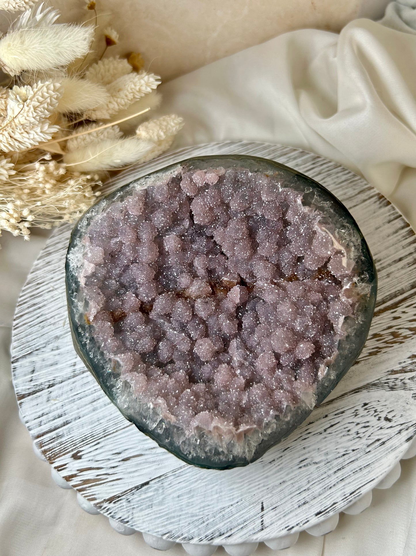 Pink Sugar Amethyst geode from Uruguay with sparkling druzy surface and soft pastel crystals, displayed on a rustic white stand beside dried florals.