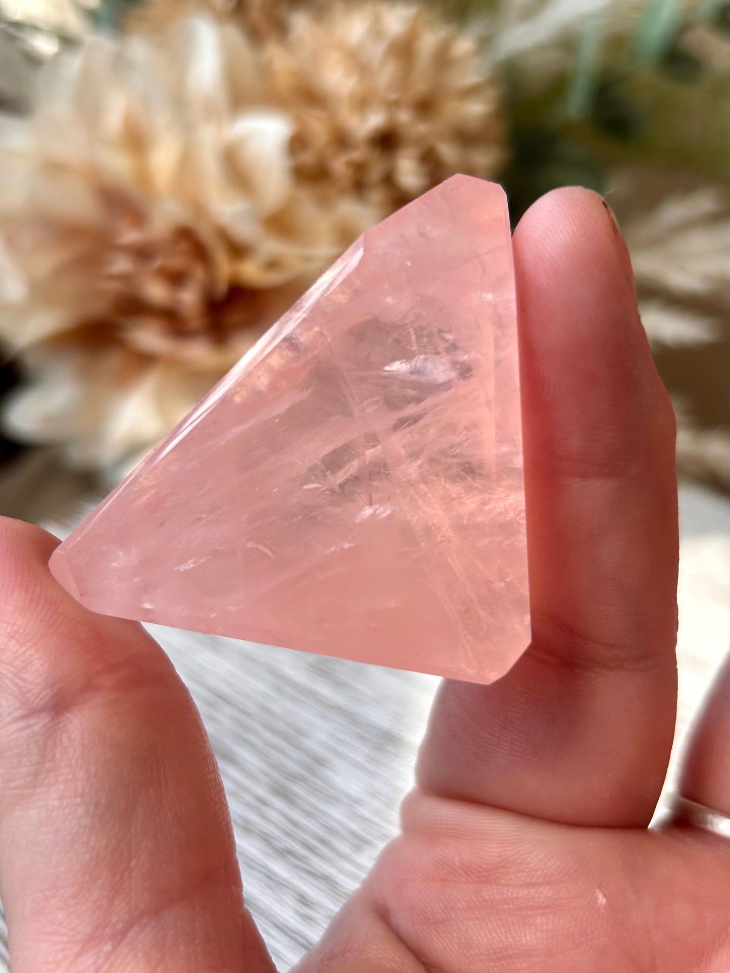 A hand holding a faceted pink crystal stone with natural inclusions, set against a soft-focus background of dried flowers.