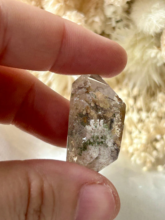 holding a Lodolite freeform crystal against an ivory satin backdrop with dried cream flowers; soft natural light highlights internal inclusions.