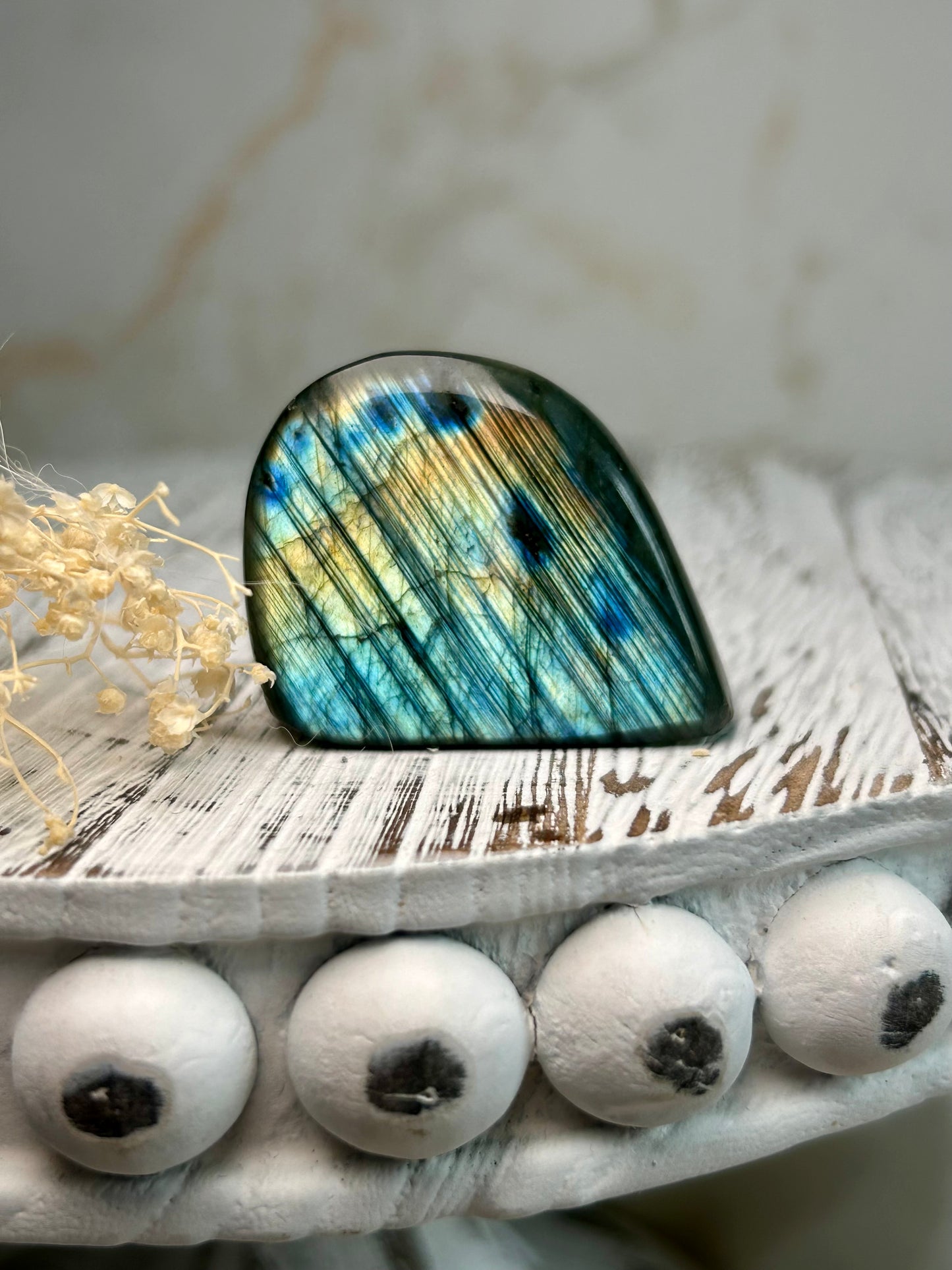 Polished labradorite freeform with vivid blue flash and iridescent surface on a distressed white wooden board with dried cream flowers