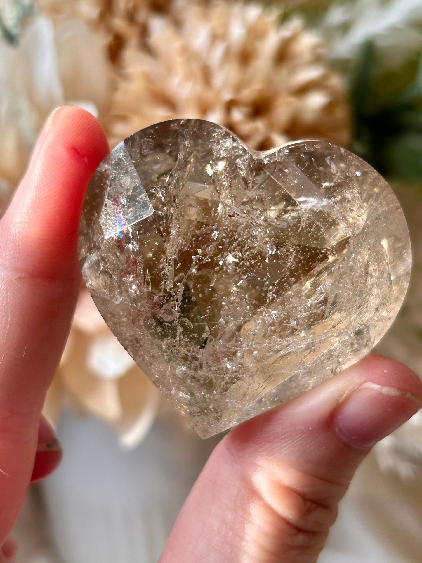A heart-shaped clear quartz crystal held between fingers, showcasing its natural internal inclusions and facets against a soft, blurred background of dried flowers.