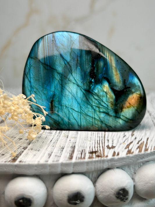 Polished labradorite freeform with vivid blue flash and iridescent surface on a distressed white wooden board with dried cream flowers.