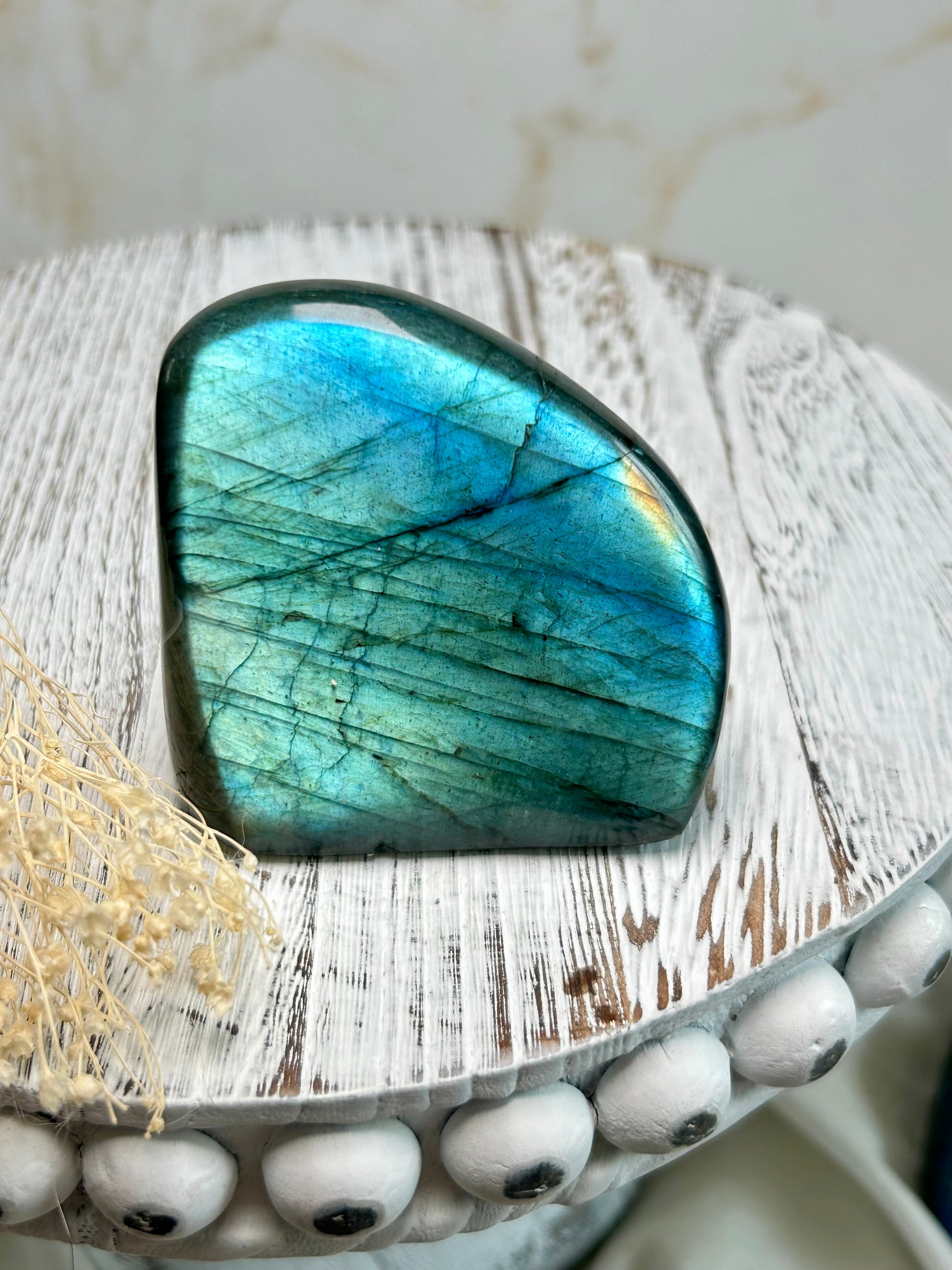 Polished labradorite freeform with vivid blue flash and iridescent surface on a distressed white wooden board with dried cream flowers