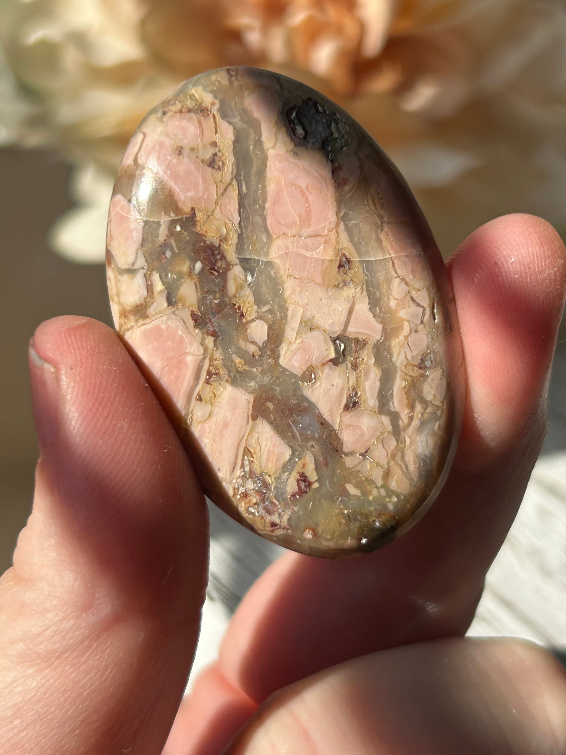 A hand holding a polished oval cabochon stone with a natural marbled pattern of pink, beige, and gray.