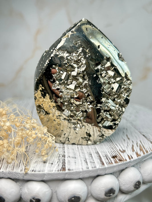 Golden pyrite flame crystal polished into flame shape, shimmering metallic surface displayed on a distressed wooden board with cream dried flowers.