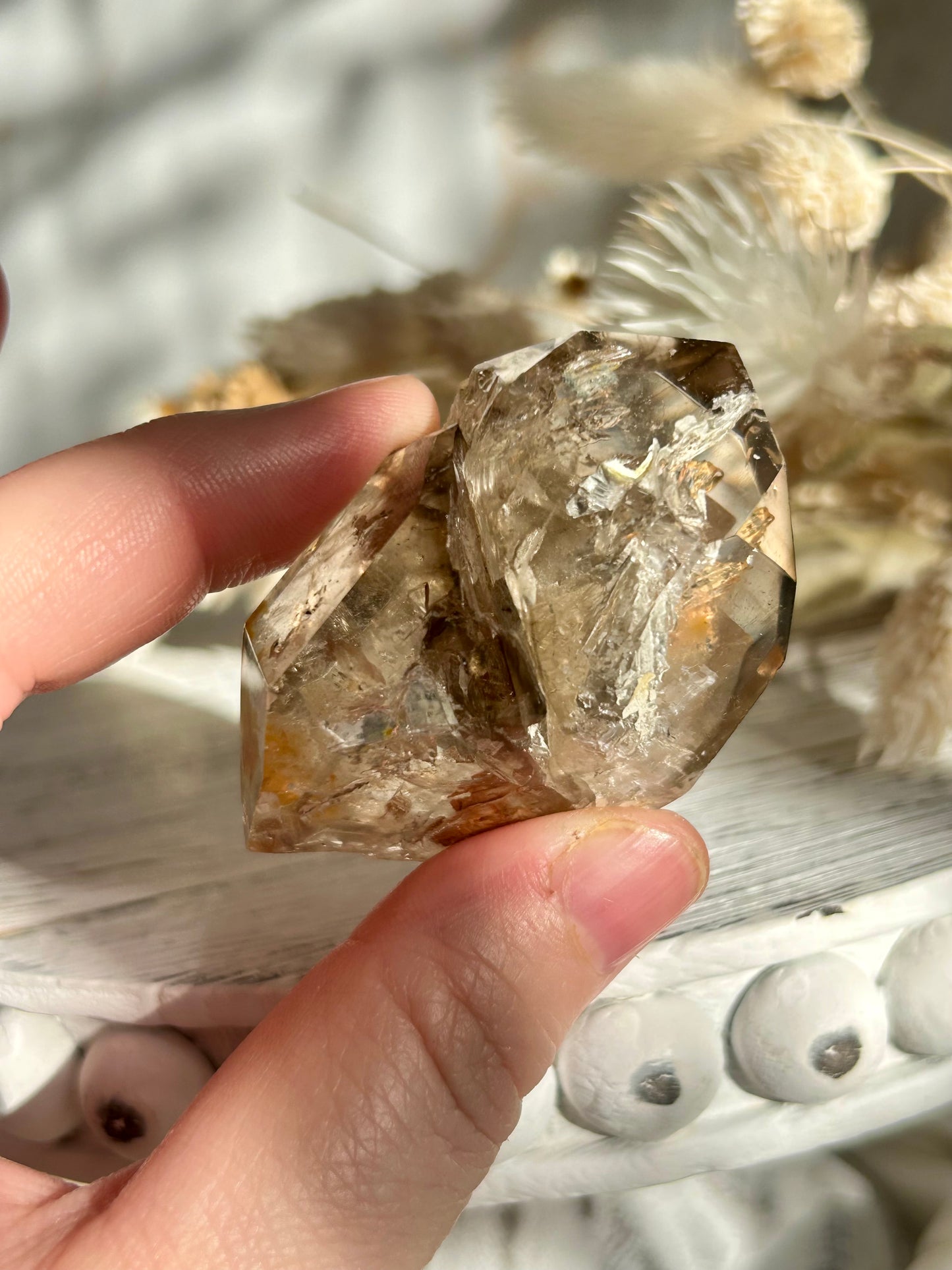 Golden Elestial Quartz crystal tower with visible layered terminations and internal inclusions, polished to a high shine. Photographed in warm natural light with soft cream dried florals in the background.