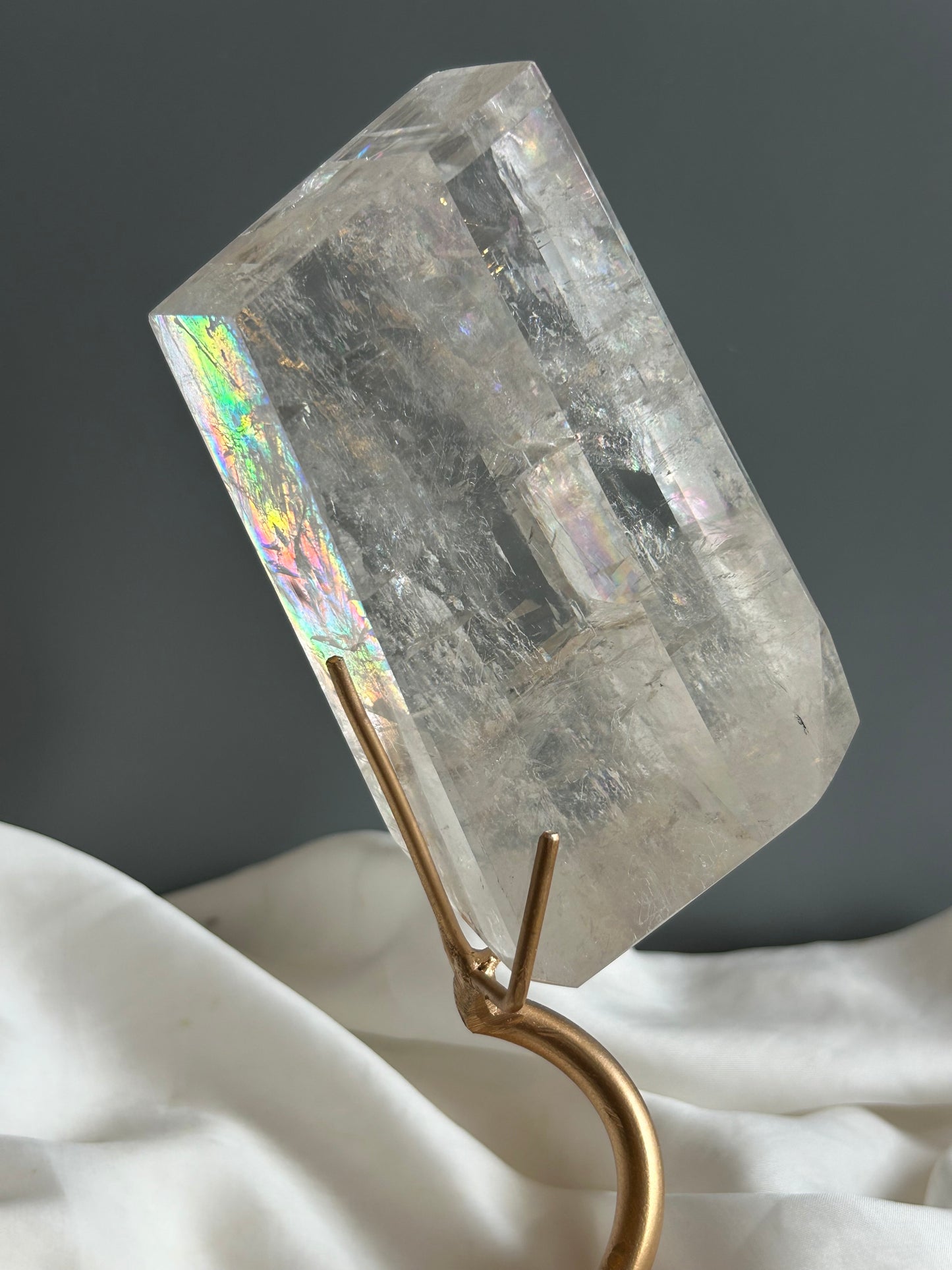 Clear Optical Calcite crystal on gold display stand showing strong light refraction and internal rainbows against a soft white fabric background.
