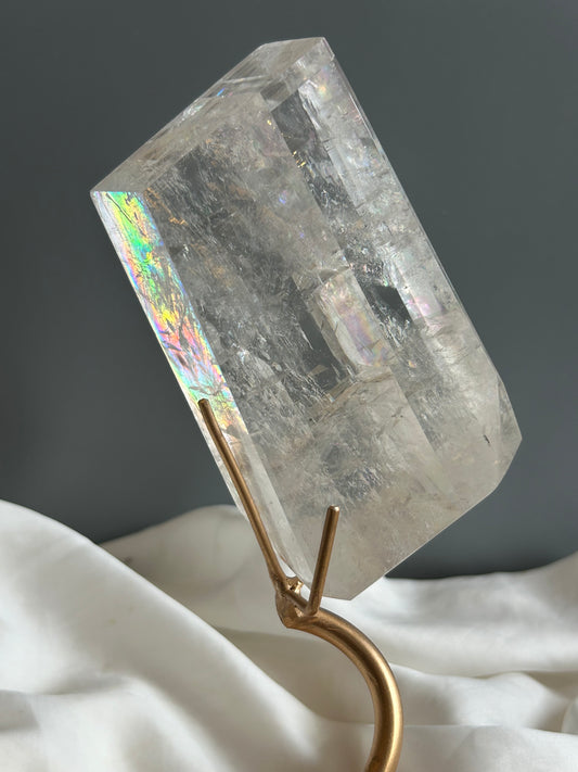Clear Optical Calcite crystal on gold display stand showing strong light refraction and internal rainbows against a soft white fabric background.