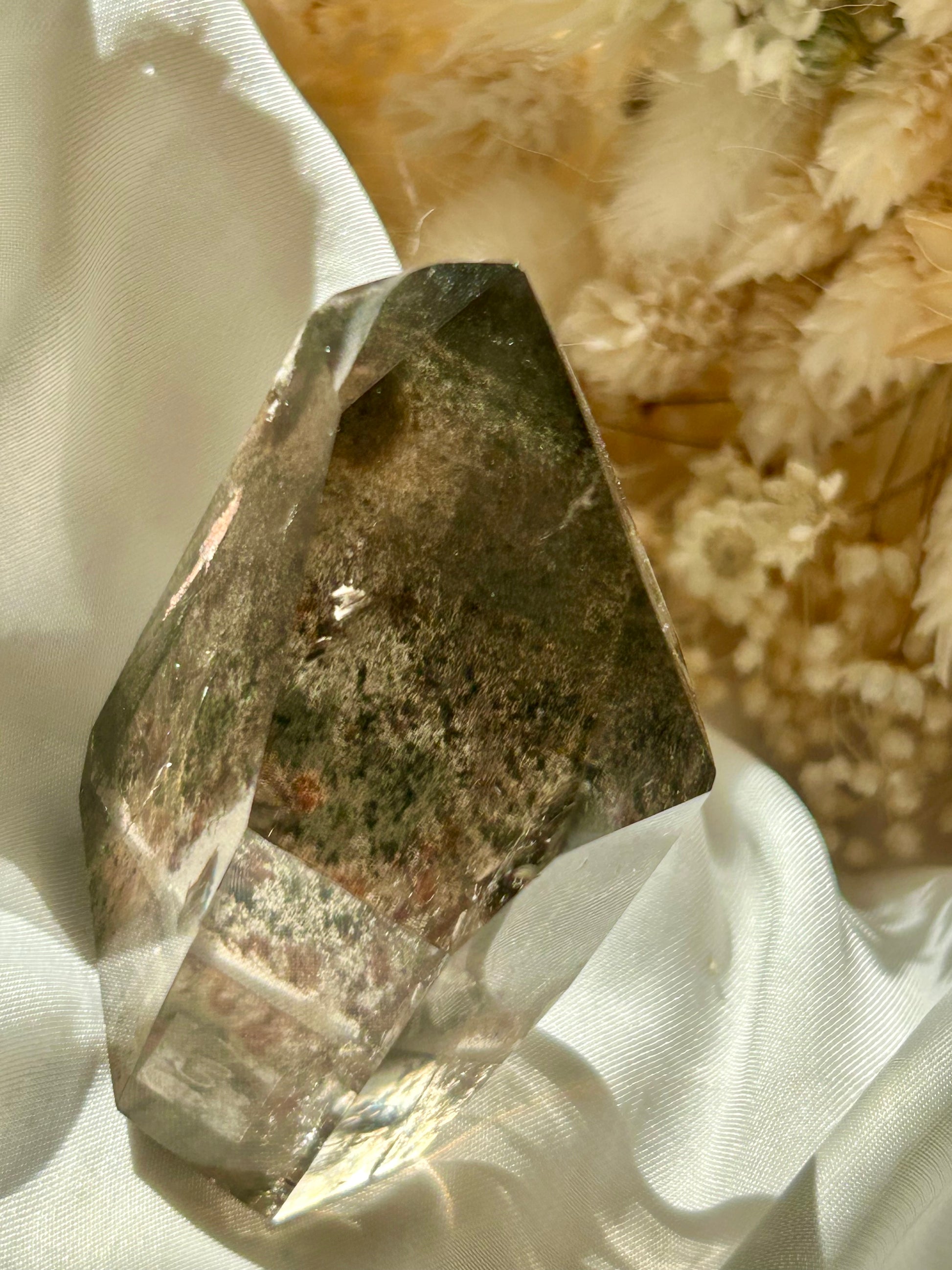 Smoky Quartz tower with chlorite inclusions resting on ivory satin, catching golden sunlight with earthy internal reflections.