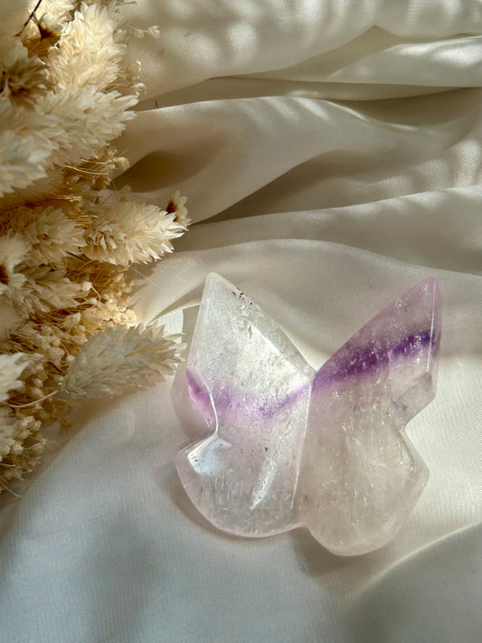 Phantom Amethyst butterfly crystal displayed on ivory satin with cream dried flower accents — gentle feminine energy and transformation stone.