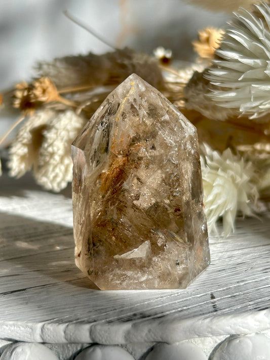 Golden Elestial Quartz crystal tower with visible layered terminations and internal inclusions, polished to a high shine. Photographed in warm natural light with soft cream dried florals in the background.