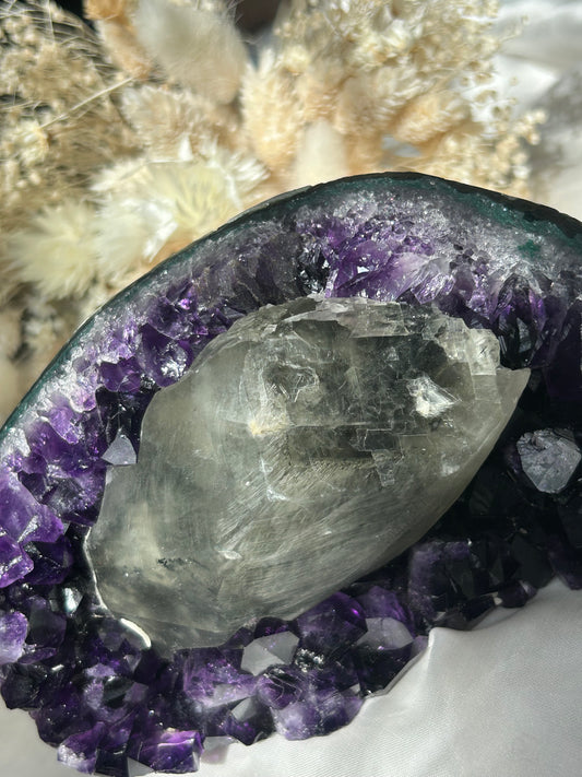 Uruguayan Amethyst geode with large calcite inclusion, deep purple crystal points and polished face, displayed with dried flowers
