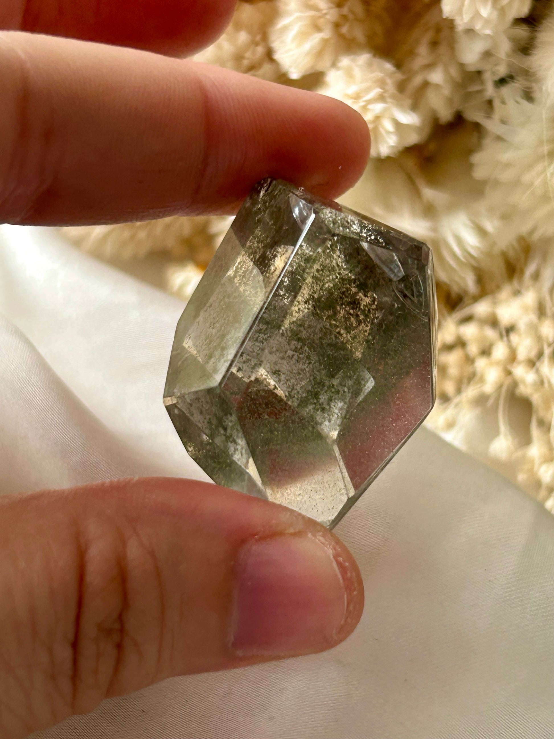 holding a Lodolite freeform crystal against an ivory satin backdrop with dried cream flowers; soft natural light highlights internal inclusions.