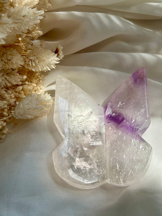 Phantom Amethyst butterfly crystal displayed on ivory satin with cream dried flower accents — gentle feminine energy and transformation stone.
