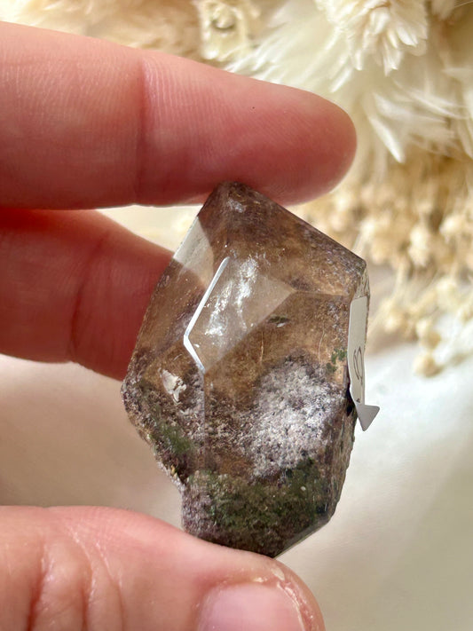 holding a Lodolite freeform crystal against an ivory satin backdrop with dried cream flowers; soft natural light highlights internal inclusions.