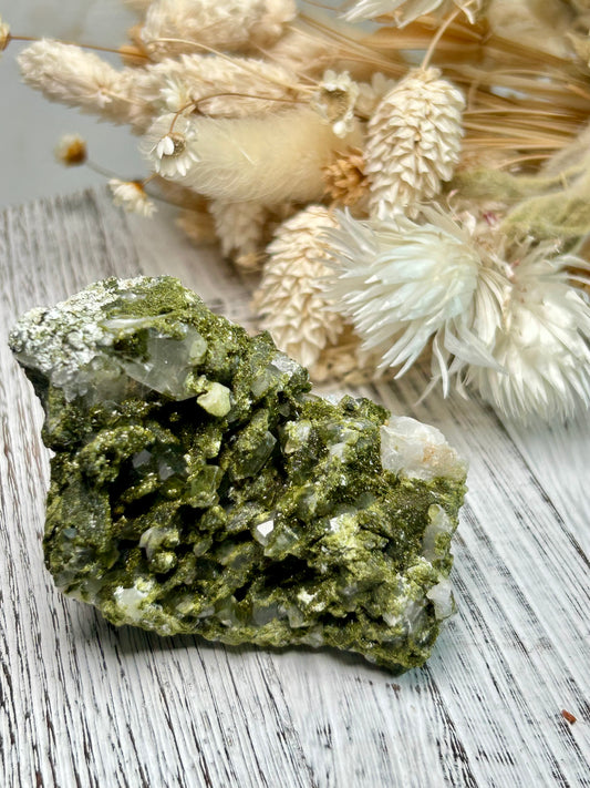 Clear quartz crystal with deep green epidote inclusions, polished freeform displayed on a distressed white wooden board with dried cream flowers 