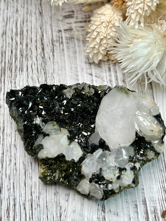 Clear quartz crystal with deep green epidote inclusions, polished freeform displayed on a distressed white wooden board with dried cream flowers 
