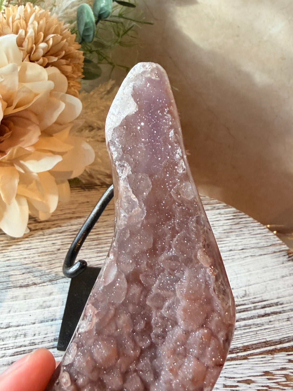A polished natural crystal or geode specimen with a dusty lavender-purple interior and white crystalline outer edges, displayed on a small black stand. A human finger is visible at the bottom left holding the item, and the scene includes decorative flowers and a rustic wood surface in the background.