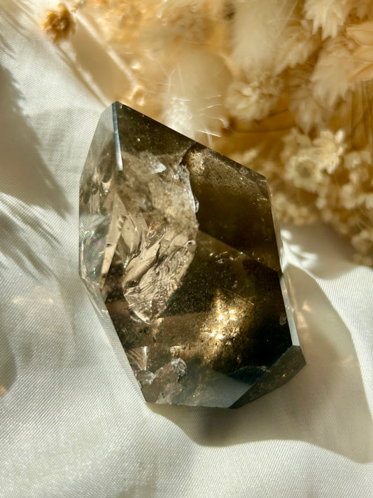 Smoky Quartz freeform with chlorite inclusions resting on ivory satin, catching golden sunlight with earthy internal reflections.