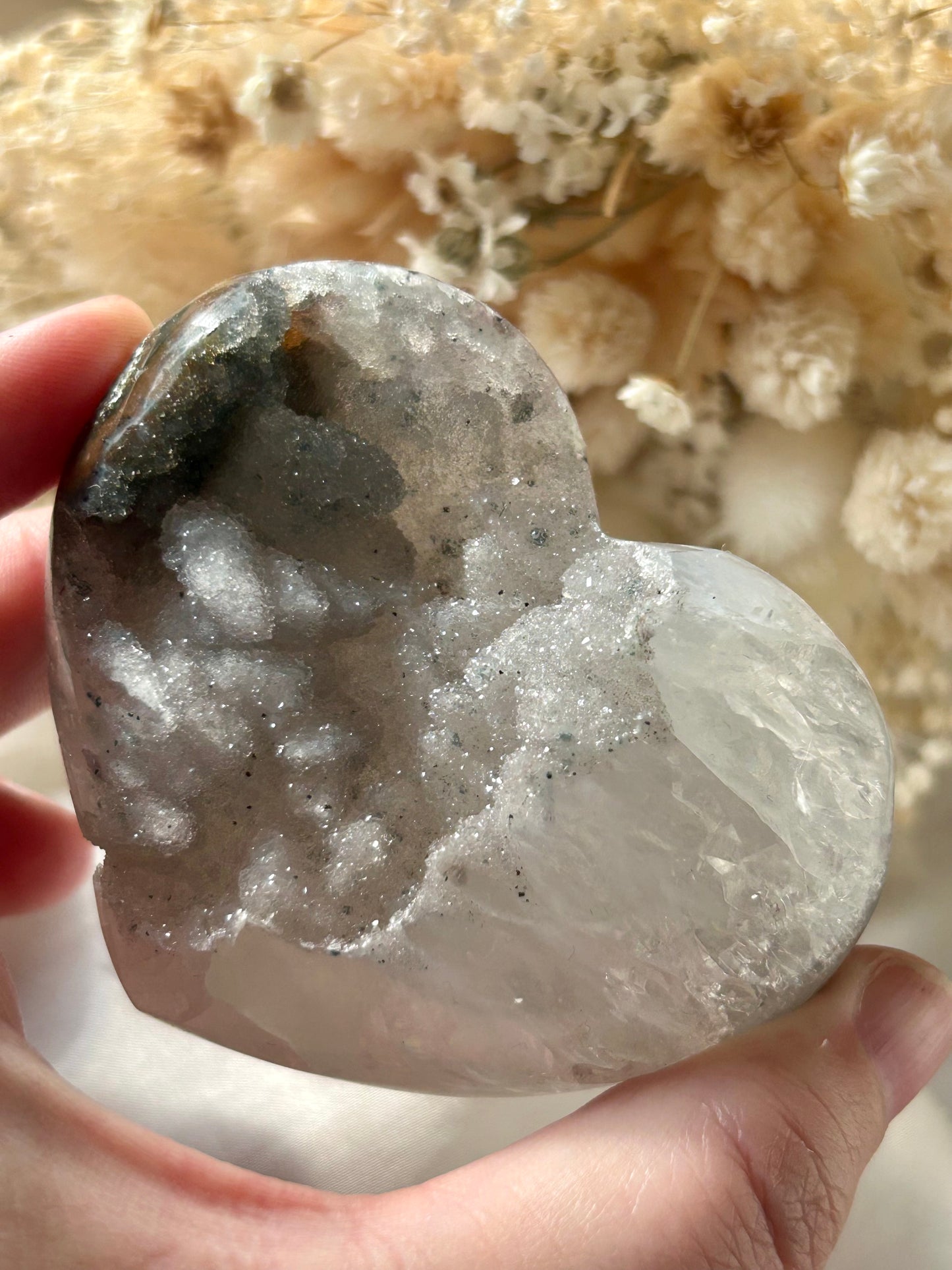 Clear Quartz crystal heart with druzy sparkle and smoky mineral veiling, held in hand against soft dried flower background