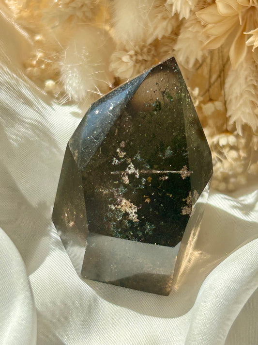 Smoky Quartz tower with chlorite inclusions resting on ivory satin, catching golden sunlight with earthy internal reflections.