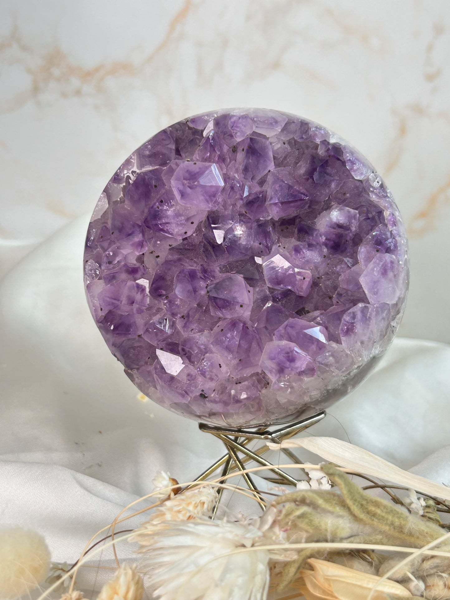 Deep lavender Amethyst druzy sphere with sparkling crystal points on a metal stand, surrounded by dried florals on white fabric.