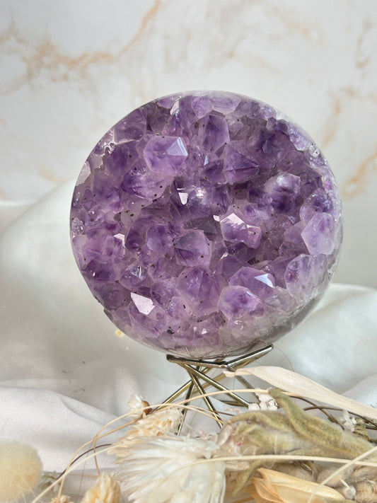 Deep lavender Amethyst druzy sphere with sparkling crystal points on a metal stand, surrounded by dried florals on white fabric.