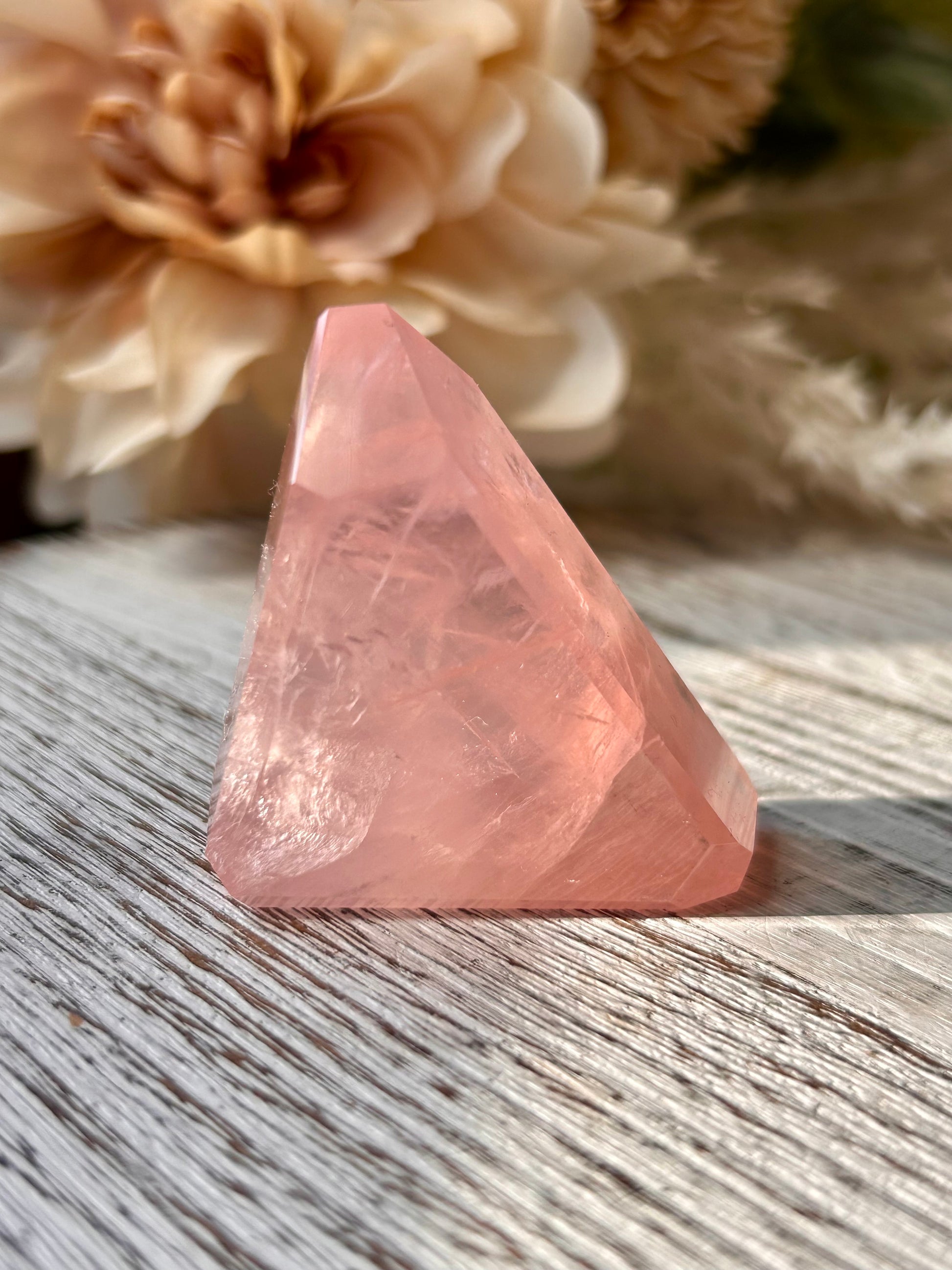 A faceted pink crystal pyramid resting on a weathered gray wooden surface, with soft beige flowers blurred in the background.