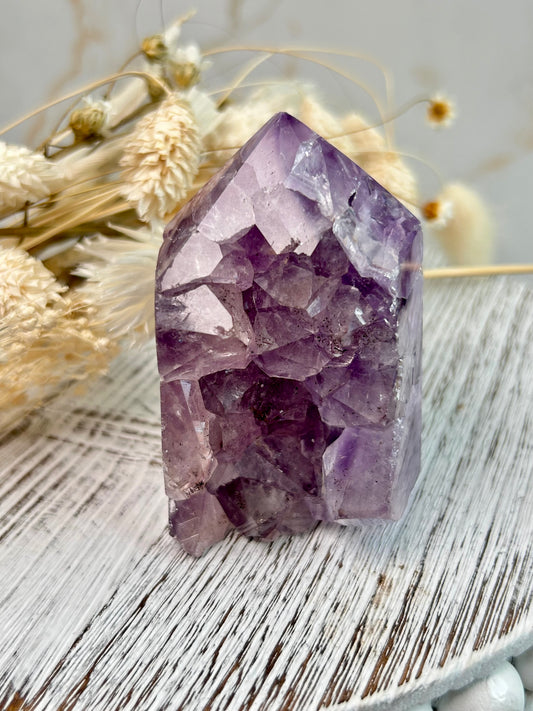 Brazilian Amethyst tower with natural internal crystal clusters, photographed on a textured white wooden surface with dried florals in the background.