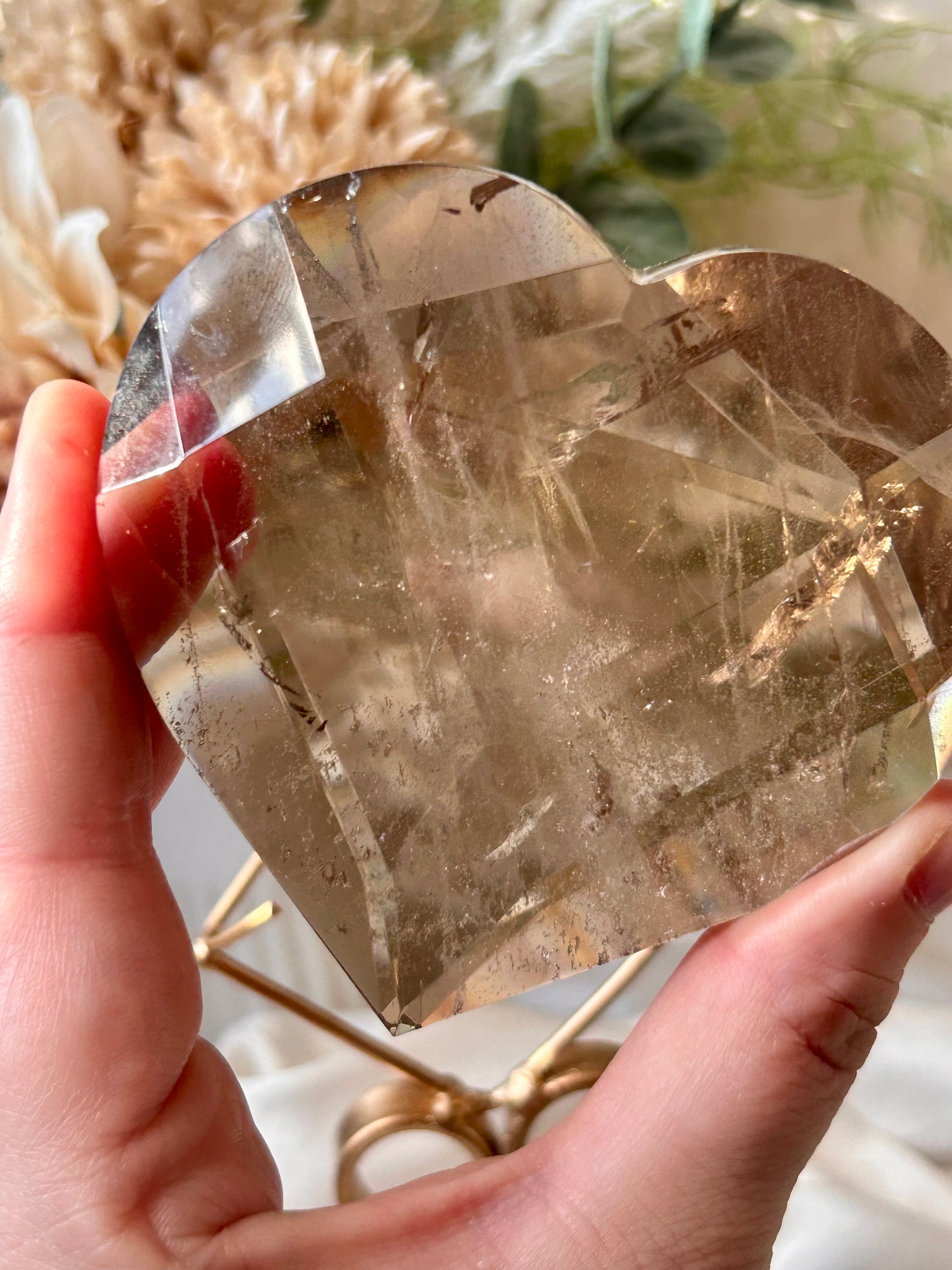 A hand holding a heart-shaped smoky quartz crystal cluster, showing its natural internal structure and translucent golden-brown color, with a blurred background of dried flowers and greenery.