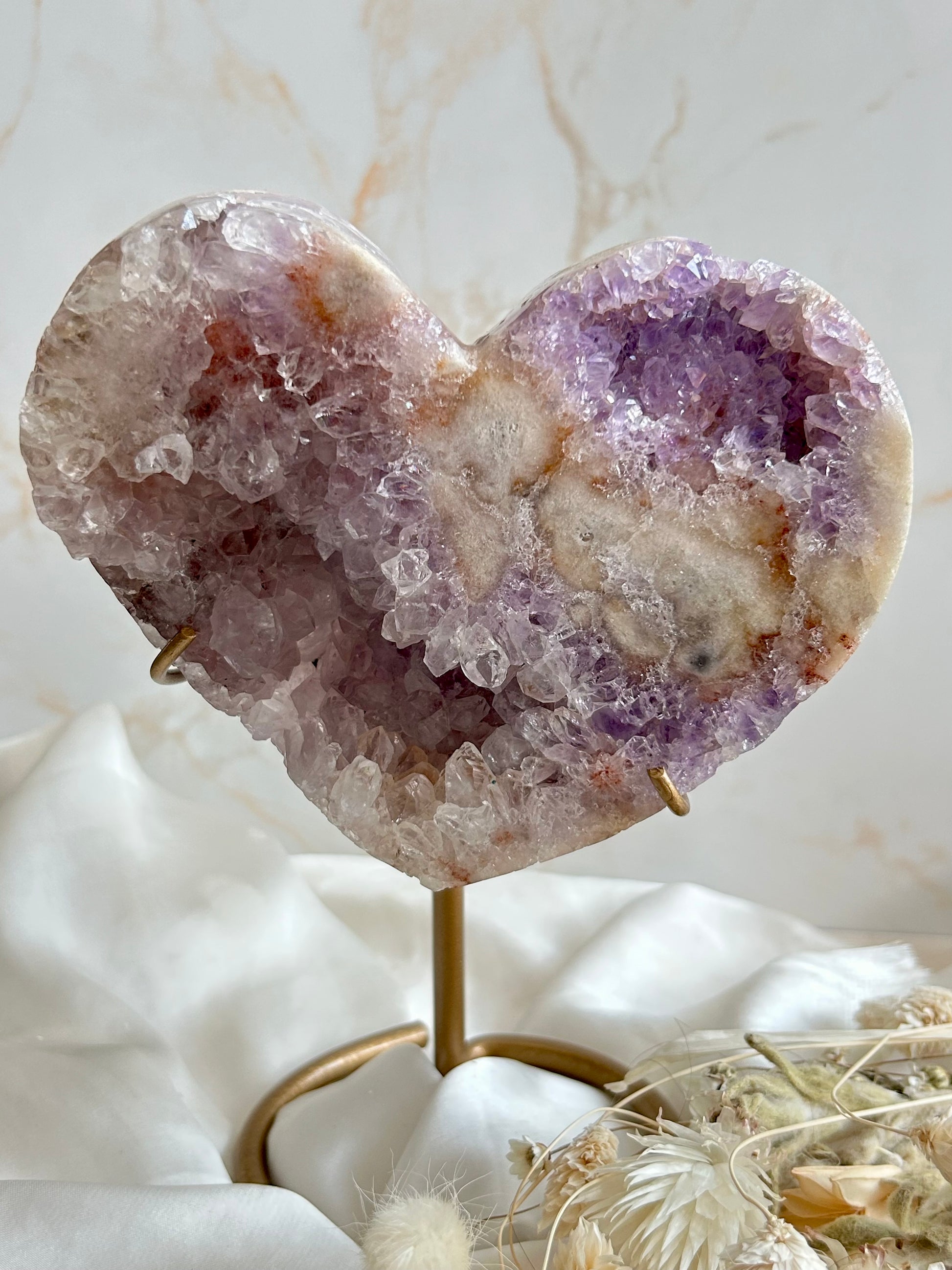 Amethyst druzy heart with lavender, clear, and peach mineral tones, held in hand. Sparkling crystal clusters and natural formations visible across the surface