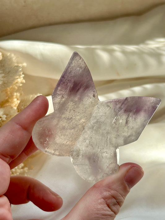 Phantom Amethyst butterfly crystal displayed on ivory satin with cream dried flower accents — gentle feminine energy and transformation stone.
