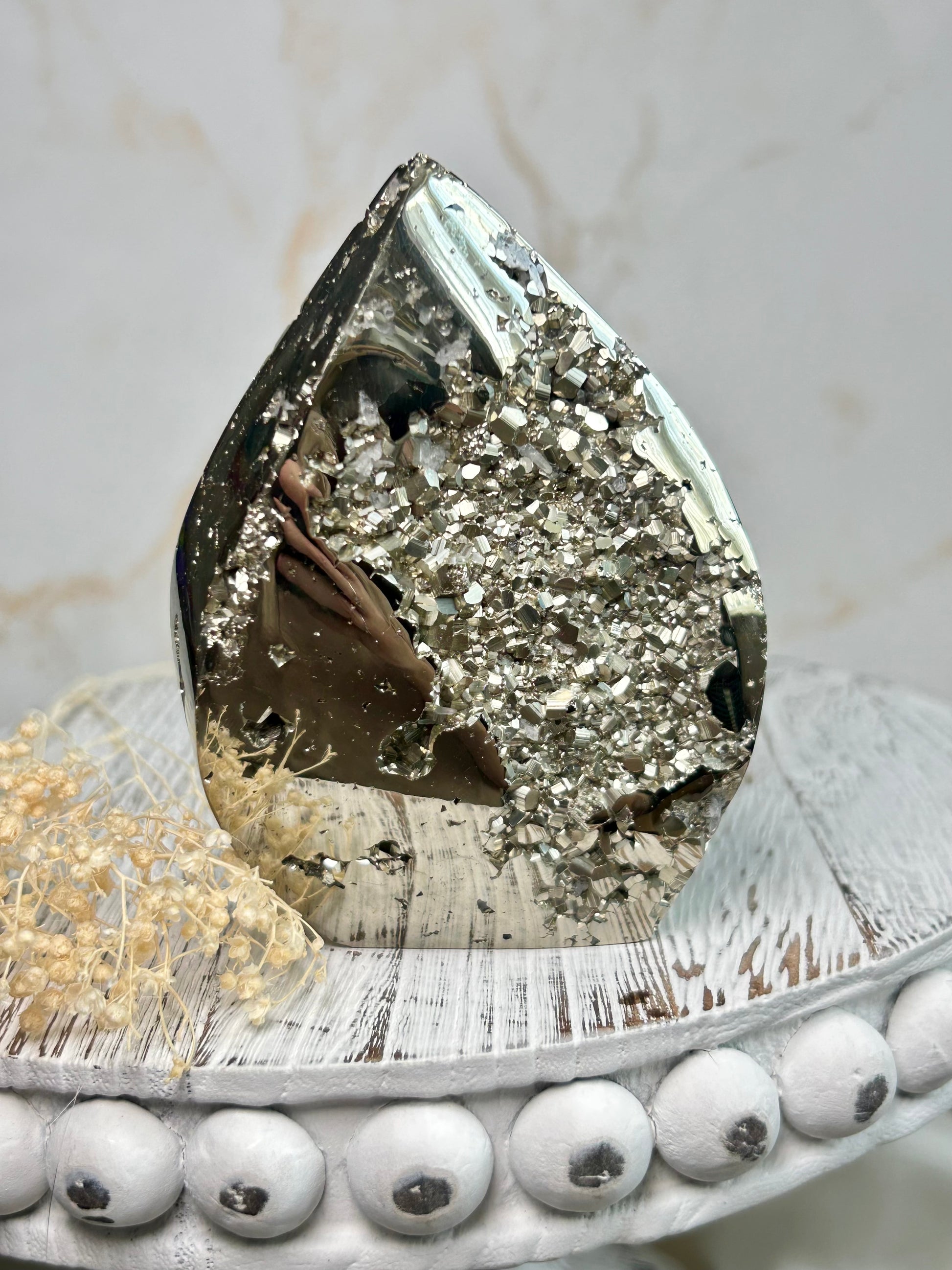 Golden pyrite flame crystal polished into flame shape, shimmering metallic surface displayed on a distressed wooden board with cream dried flowers.