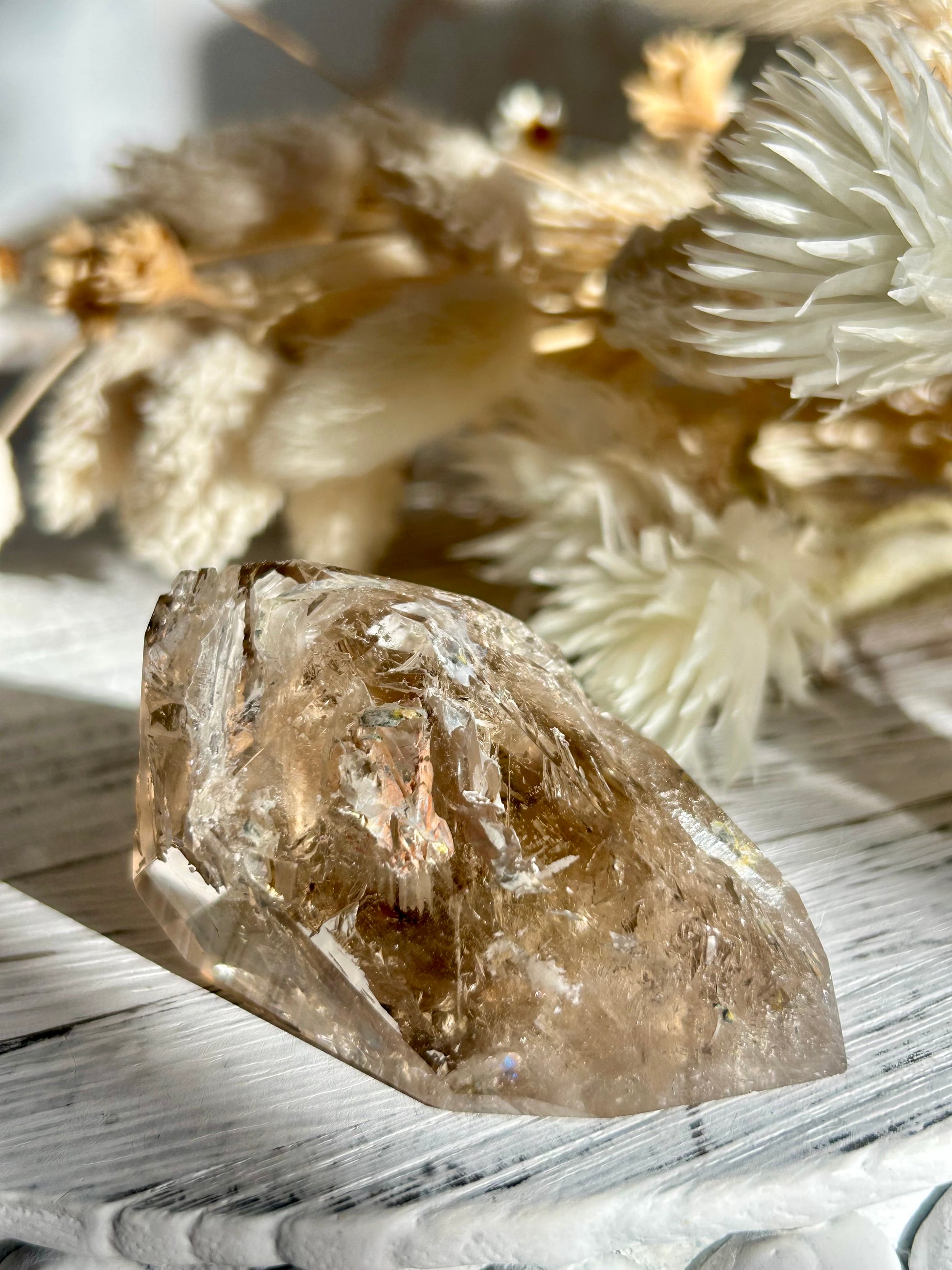 Golden Elestial Quartz crystal tower with visible layered terminations and internal inclusions, polished to a high shine. Photographed in warm natural light with soft cream dried florals in the background.