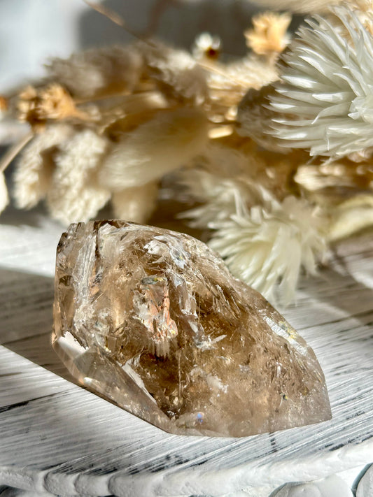 Golden Elestial Quartz crystal tower with visible layered terminations and internal inclusions, polished to a high shine. Photographed in warm natural light with soft cream dried florals in the background.