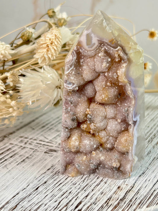 A polished Brazilian Agate tower with soft caramel and lilac bubble-like cluster formations, photographed against a neutral background with dried florals.
