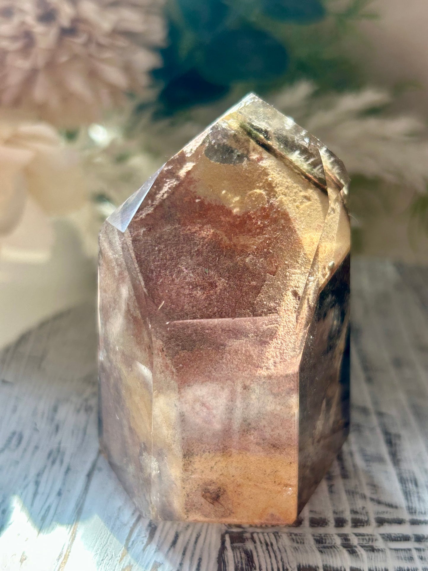 A natural faceted crystal tower with layered translucent bands of brown, gold, and dark smoky tones, displayed on a rustic white wooden surface with soft blurred dried flowers in the background.