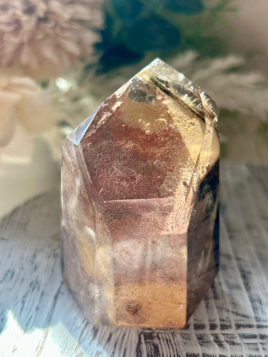 A natural faceted crystal tower with layered translucent bands of brown, gold, and dark smoky tones, displayed on a rustic white wooden surface with soft blurred dried flowers in the background.