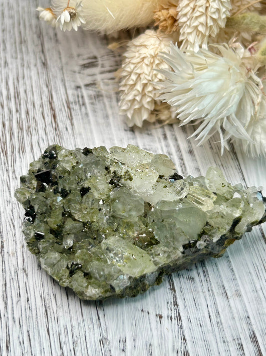 Clear quartz crystal with deep green epidote inclusions, polished freeform displayed on a distressed white wooden board with dried cream flowers 