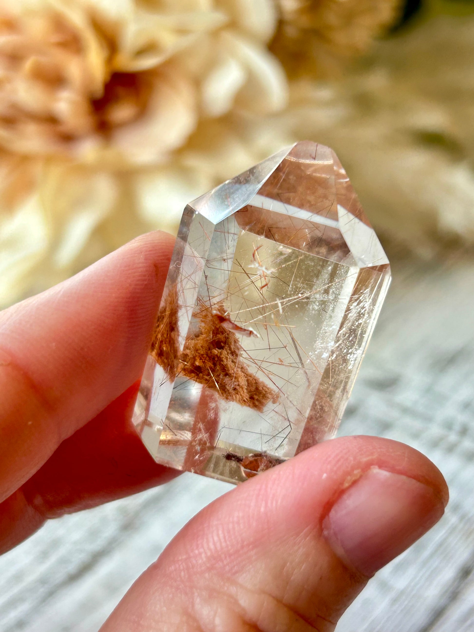 A hand holding a clear pointed crystal with visible brown and metallic inclusions, photographed close-up against a softly blurred cream floral background.