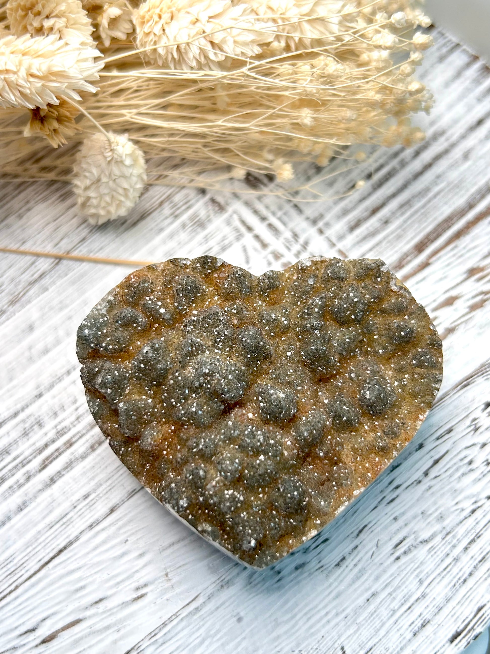 Sugar Amethyst heart from Uruguay with golden druzy surface and soft sparkling texture, displayed on a rustic white surface beside dried florals.