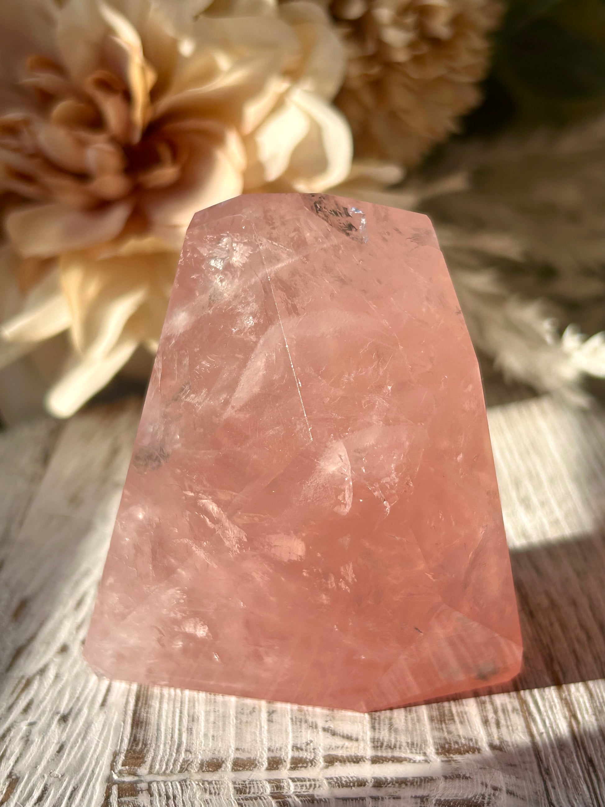 A faceted pink quartz crystal cluster, likely a rose quartz point, resting on a rustic wooden surface with soft-focus dried flowers in the background.