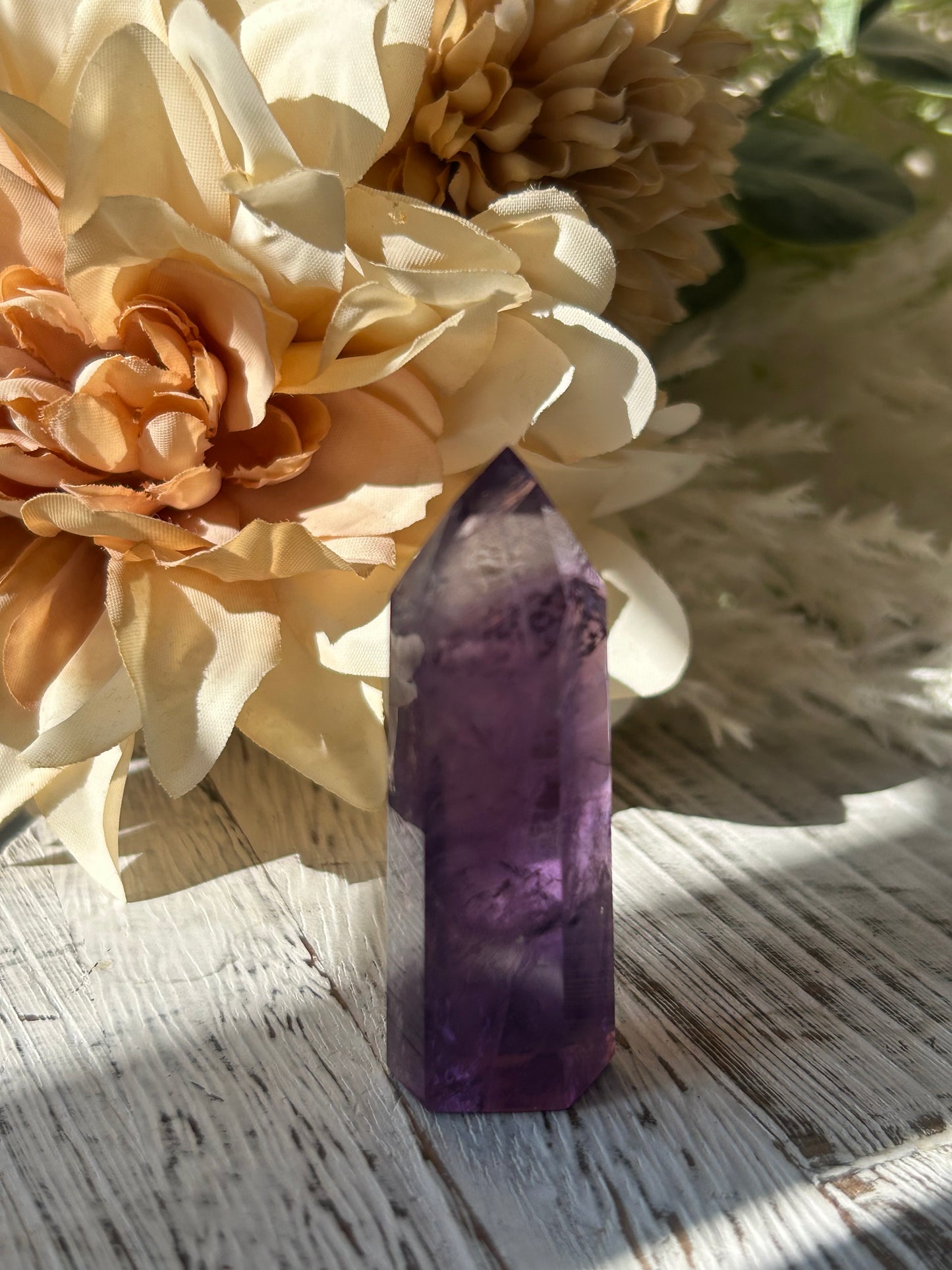 A polished purple amethyst crystal point resting on a rustic white wooden surface surrounded by cream and peach dried flowers.
