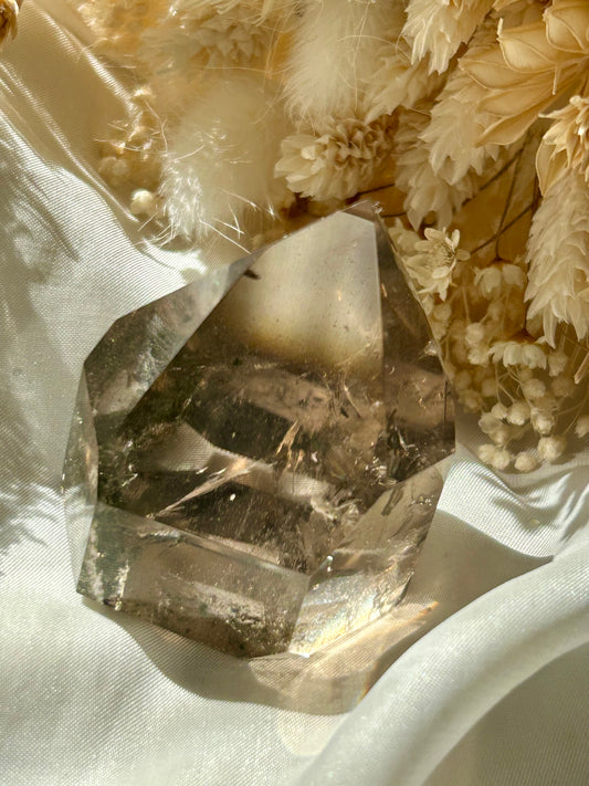 Smoky Quartz tower with chlorite inclusions resting on ivory satin, catching golden sunlight with earthy internal reflections.