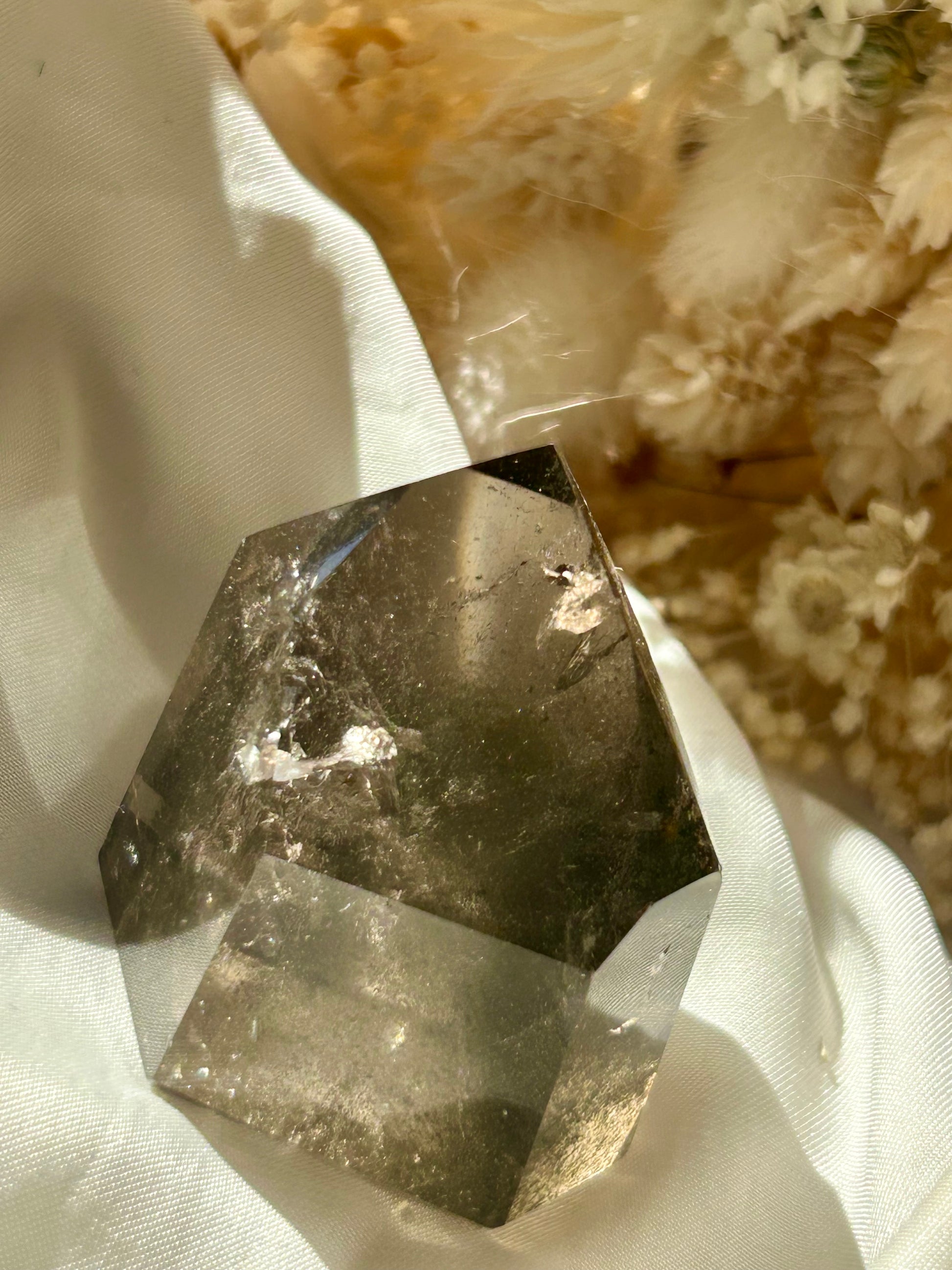 Smoky Quartz tower with chlorite inclusions resting on ivory satin, catching golden sunlight with earthy internal reflections.