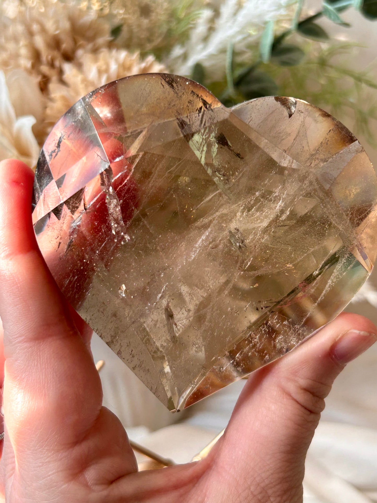 A heart-shaped clear crystal gemstone held in a person's hand, showing internal inclusions and facets, with a soft-focus background of dried flowers and greenery.